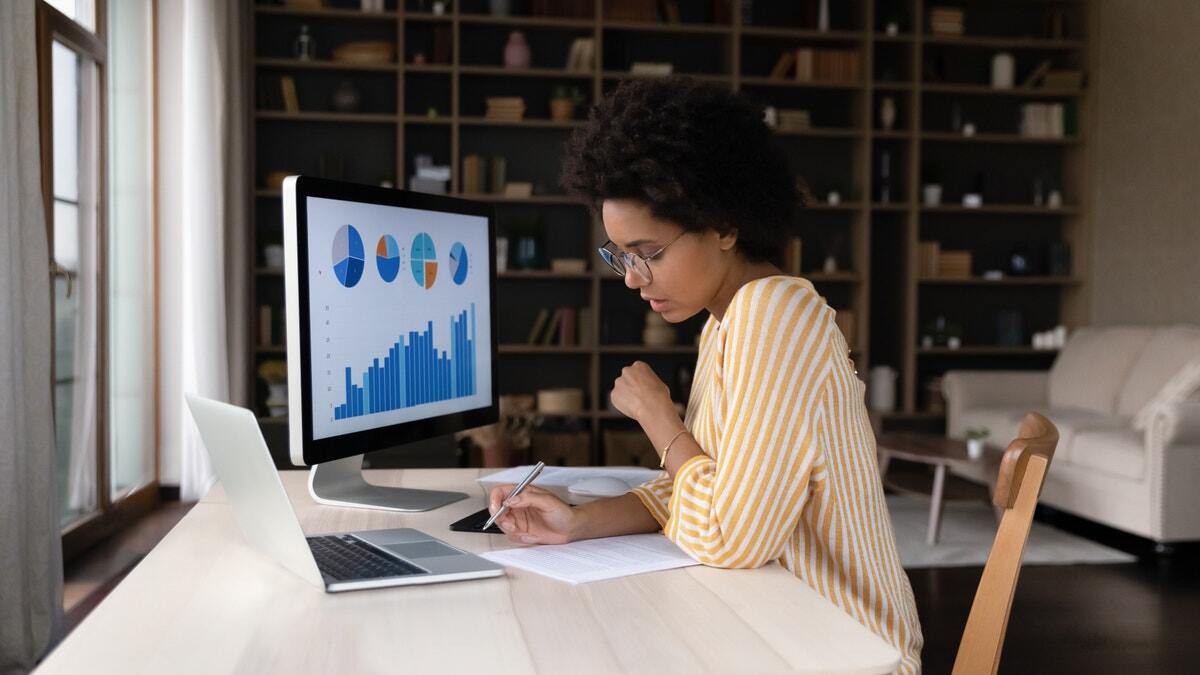 woman sitting at desk looking down at laptop with monitor